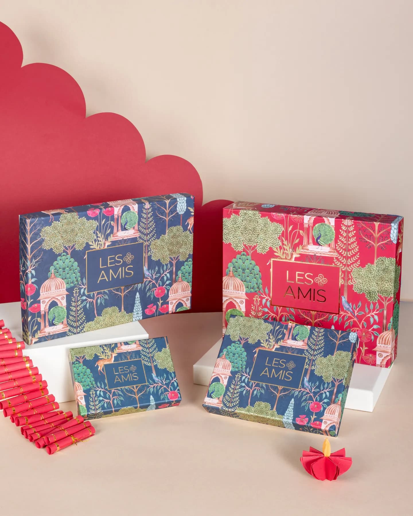 Les Amis Diwali hamper boxes in red and navy, illustrated with peacocks, pavilions and trees, on a red scalloped backdrop