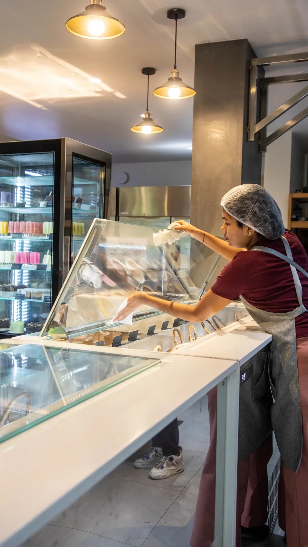 A team member polishing the glass gelato counter under warm pendant lights, colourful popsicles glowing in the display fridge behind