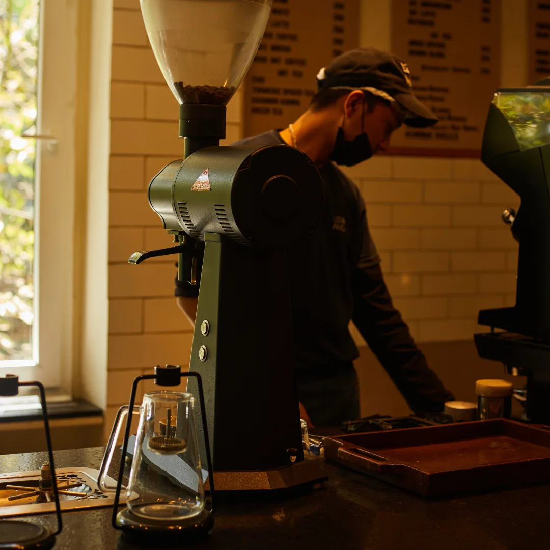 A barista working the grinder in deep golden-hour light, the menu board glowing behind