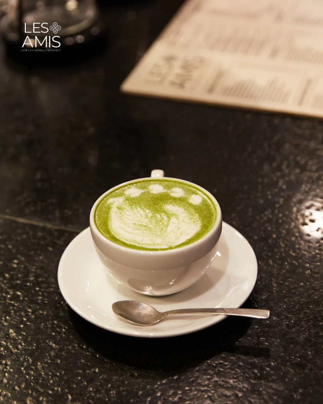 A matcha latte with delicate art on a white saucer, the café menu blurred behind