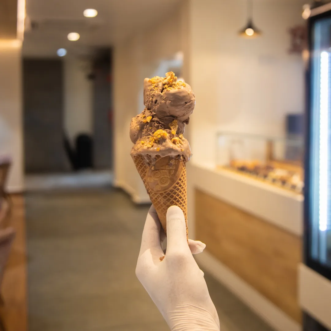 A double scoop of chocolate gelato with honeycomb crumble, held up in a waffle cone inside the café