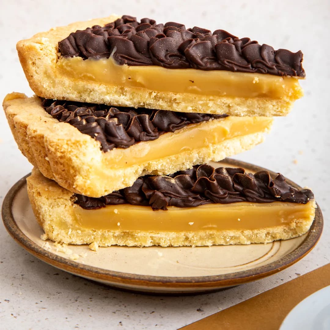 Slices of caramel chocolate tart stacked on a ceramic plate — shortbread, soft caramel and piped dark chocolate