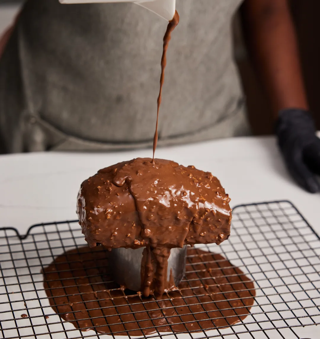 A pastry chef pouring rocher glaze over a chocolate cake on a wire rack