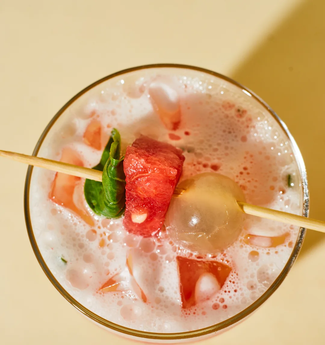 A watermelon–lychee spritzer with basil, iced and skewered, on a warm yellow table