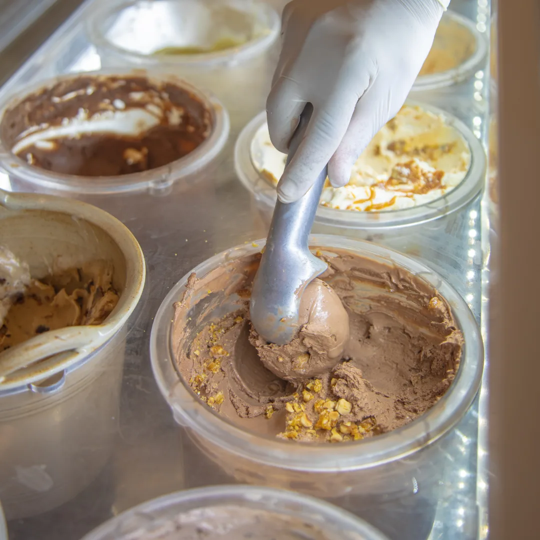 A gloved hand drawing a scoop from the display tubs, pistachio and chocolate side by side