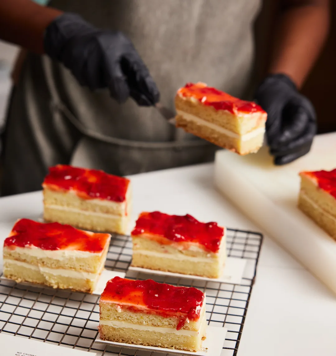 Gloved hands arranging sliced strawberries onto a pastry on a wire cooling rack